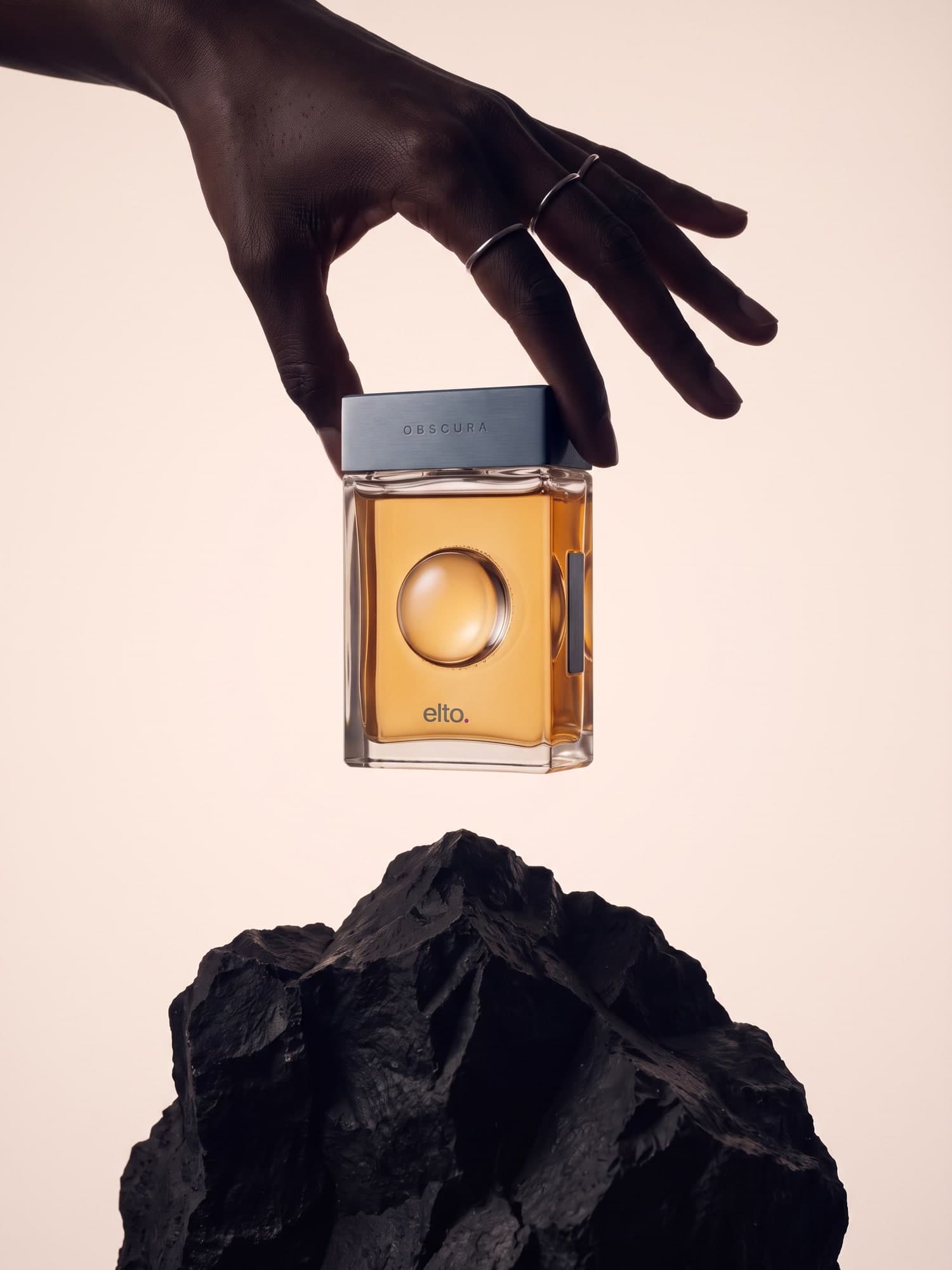 A hand with silver rings holds the amber fragrance bottle by its steel cap above a black volcanic rock